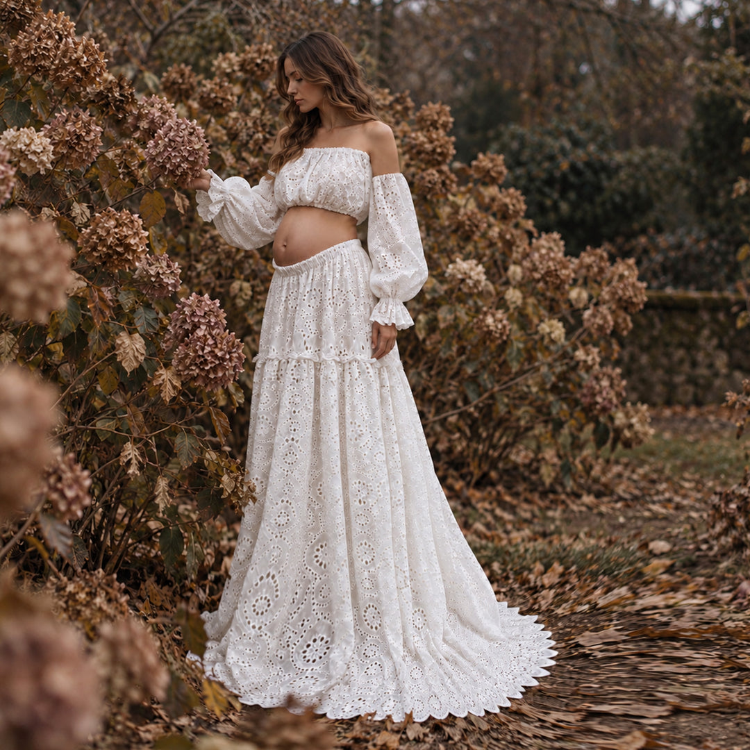 A woman wearing a white off-the-shoulder boho eyelet maxi dress stands outdoors in a garden path surrounded by autumn foliage and dried flowers.