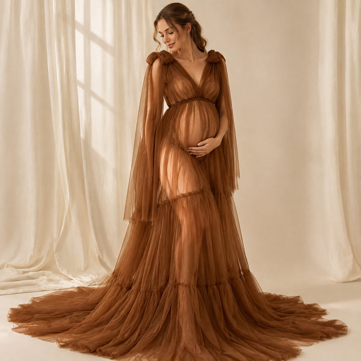 A pregnant woman in a Acaste gown, stands before white curtains, cradling her belly.