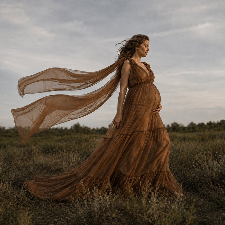 A pregnant woman stands in a grassy field wearing a flowing, sleeveless brown dress that billows in the wind. 