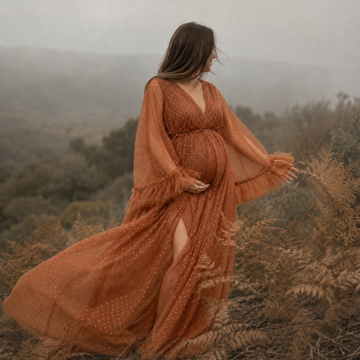 A pregnant woman stands on a hillside wearing a terracotta polka dot  tulle maternity gown with long ruffled sleeves, a V-neckline, and a high side slit.