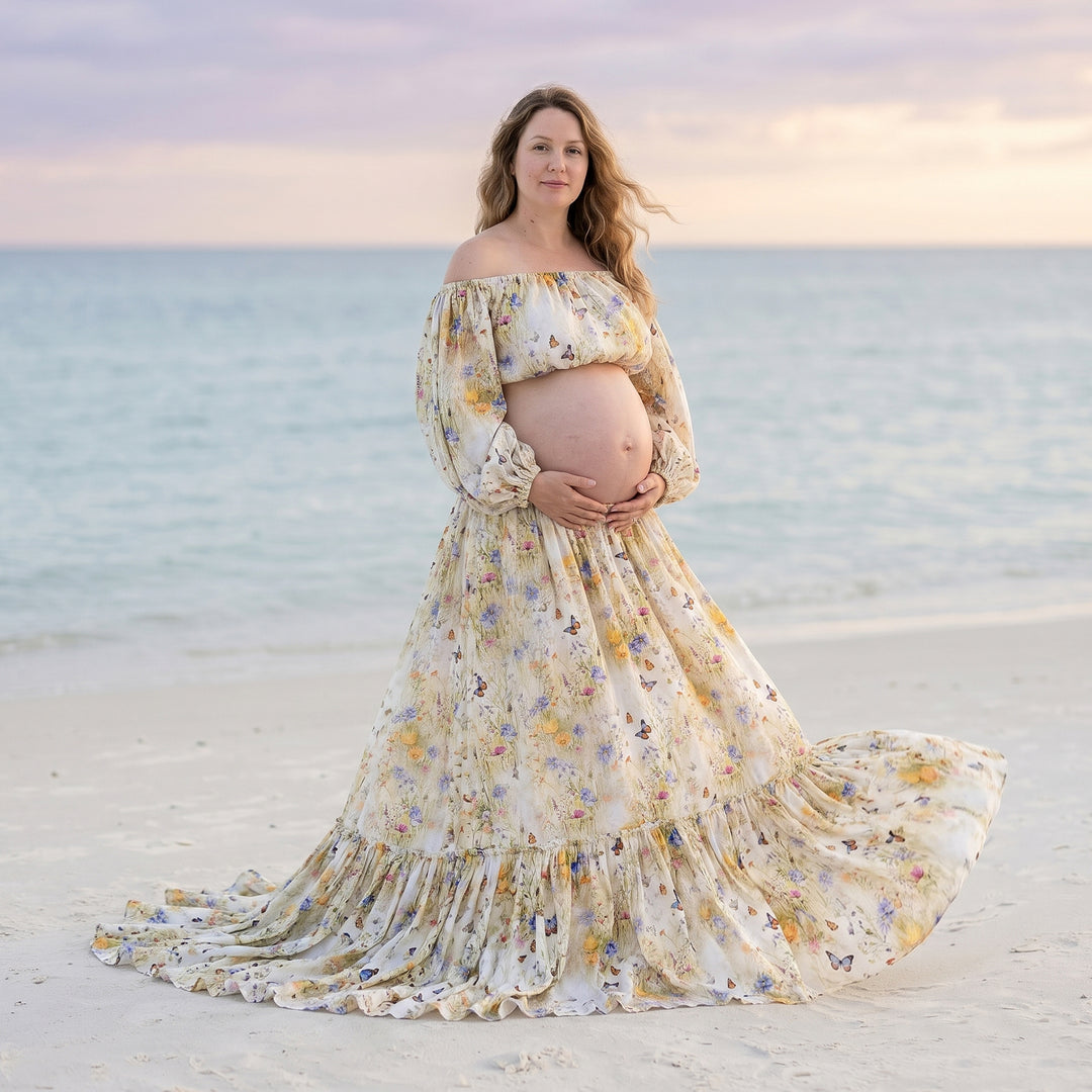A pregnant woman in a floral off-shoulder two-piece stands on the beach holding her belly.