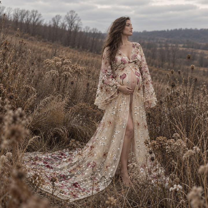 A pregnant woman stands in a dry grassy field wearing a long, sheer tulle gown with floral embroidery and sequined detailing.