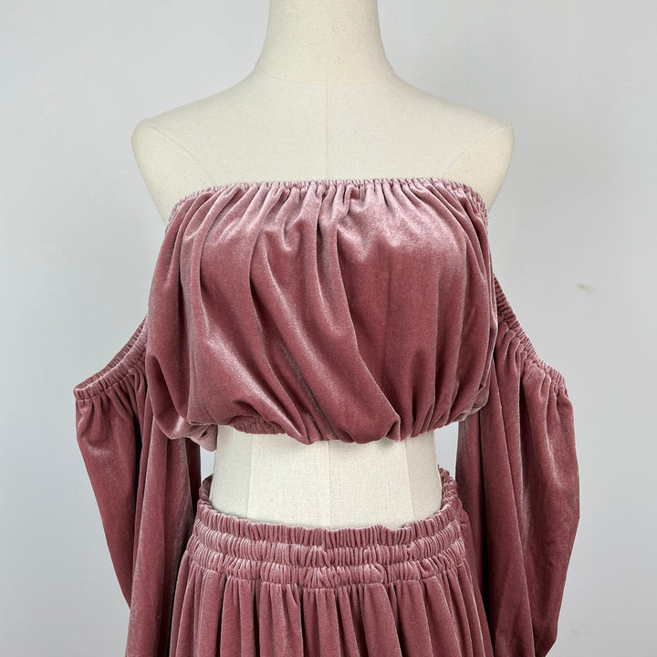 A dusty rose velvet two-piece set displayed on a cream mannequin torso. The set includes an off-the-shoulder, gathered bust crop top with long draped sleeves, and a matching gathered skirt.