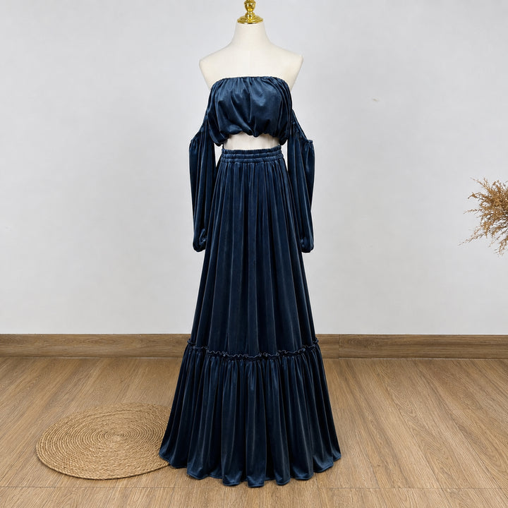A dark navy blue velvet two-piece set consisting of an off-the-shoulder crop top with long sleeves and a matching tiered maxi skirt, displayed on a mannequin in an indoor setting with a wood floor and white wall.