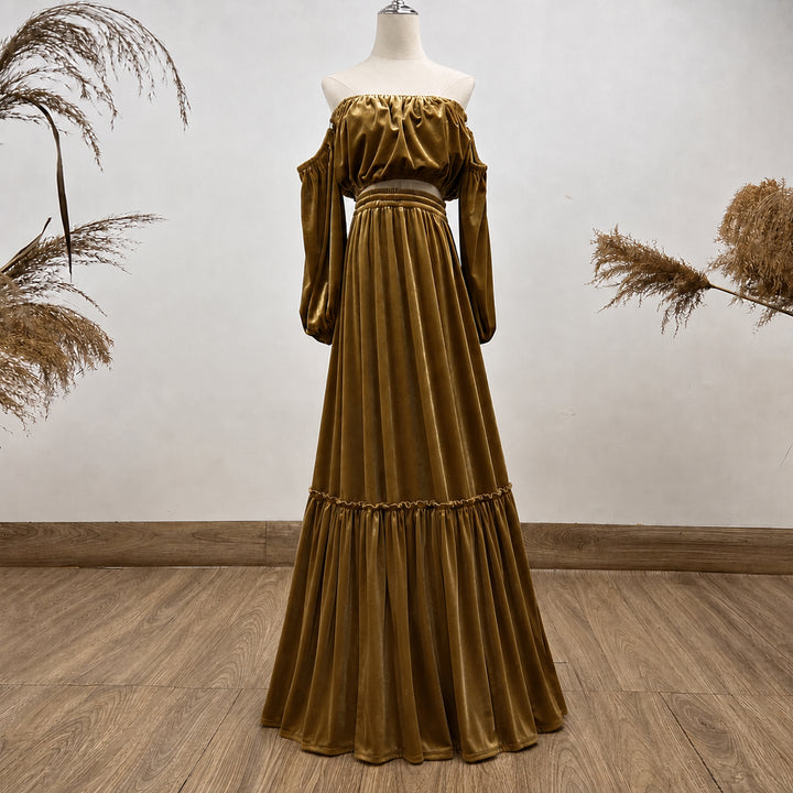 An off-the-shoulder long-sleeve maxi dress in a rich gold velvet fabric, displayed on a mannequin against a neutral wall with dried pampas grass accents.