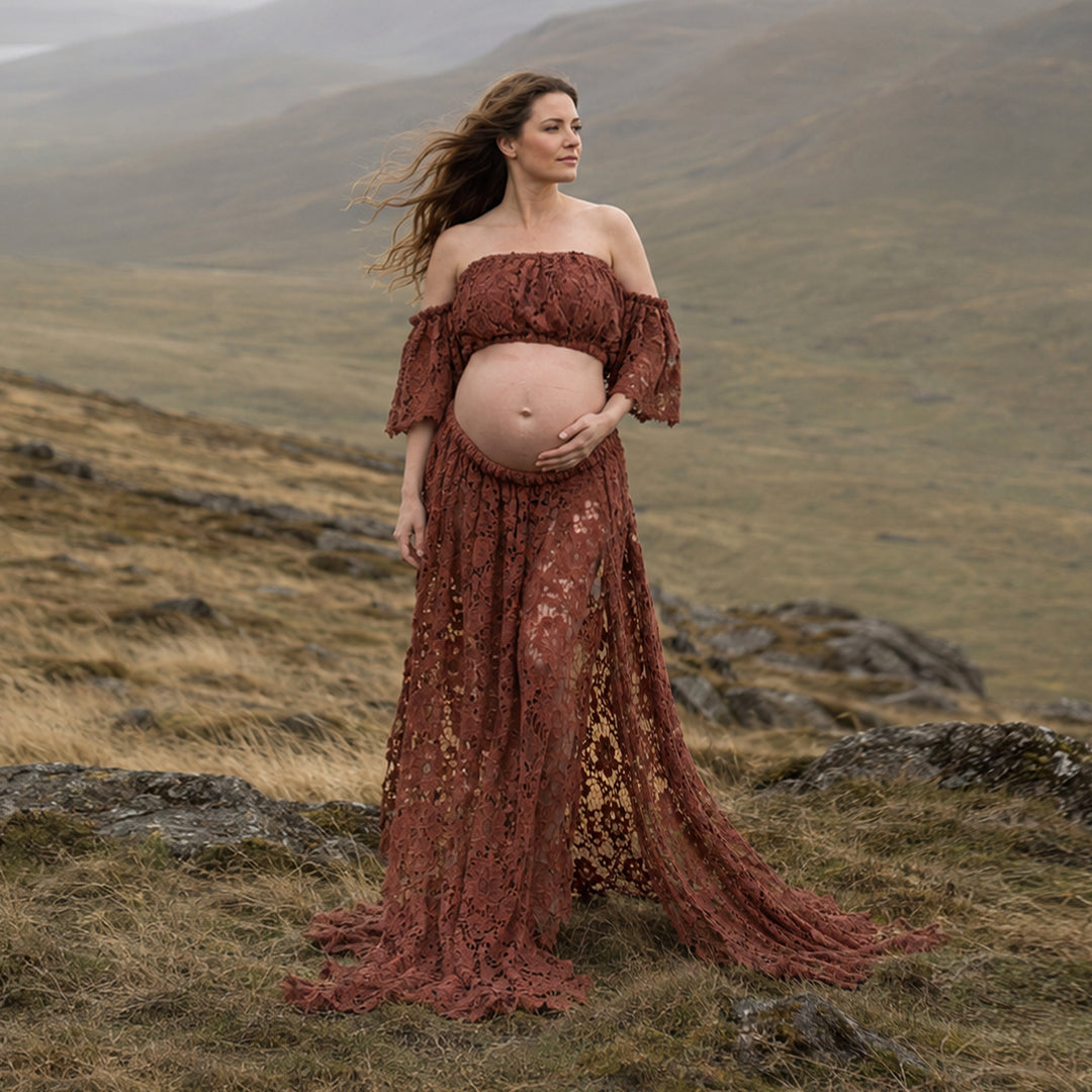 A pregnant woman stands on a grassy hillside wearing a terracotta off the shoulder lace maternity gown with ruffled sleeves and an open front skirt that reveals her baby bump and flows over the terrain.
