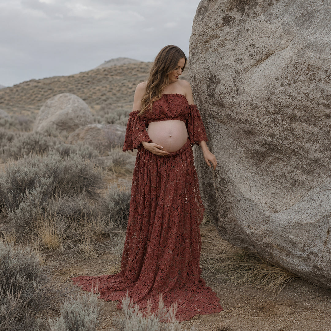 A pregnant woman stands beside a large boulder in a desert landscape wearing a Bendis terracotta off-the-shoulder lace maternity gown with ruffled sleeves and an open front skirt that reveals her baby bump.