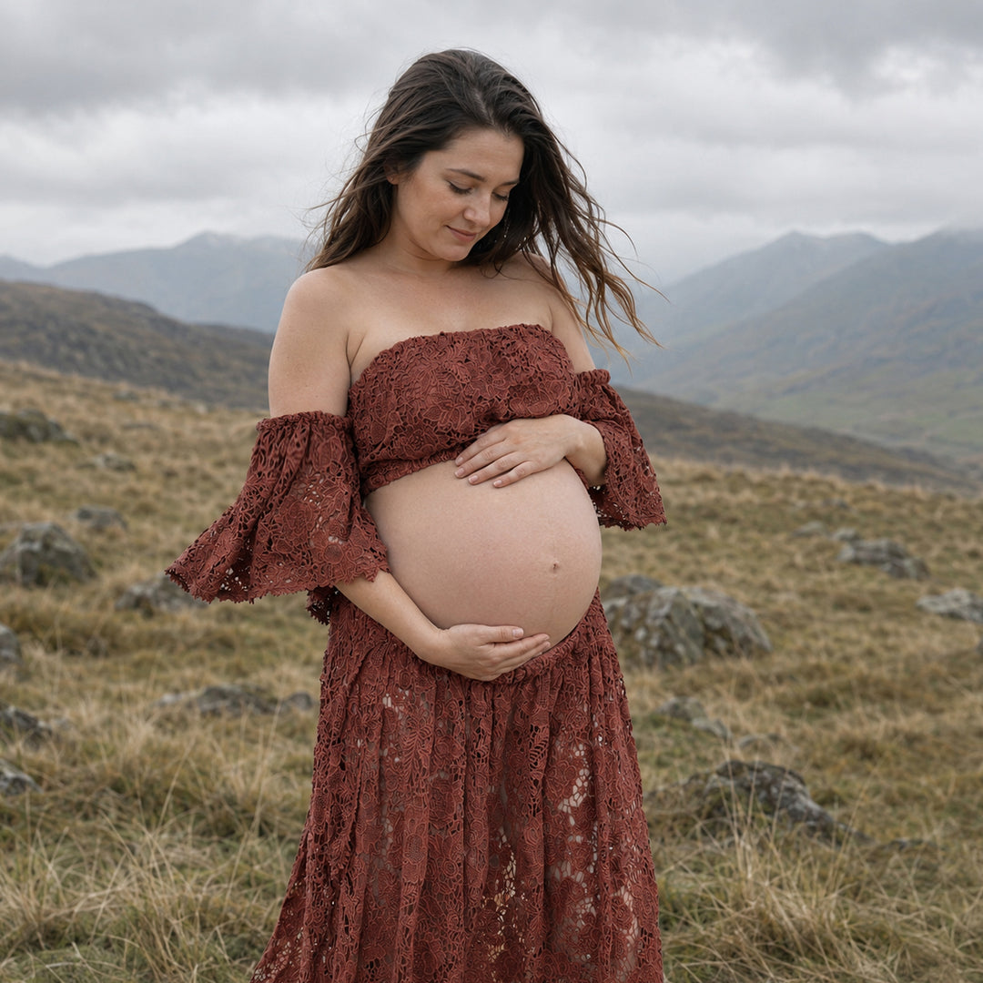 A pregnant woman stands in profile on a misty, grassy hillside wearing a Bendis terracotta two-piece lace maternity set with off-the-shoulder ruffled sleeves and a high-waisted skirt that reveals her bare baby bump against a mountain backdrop.