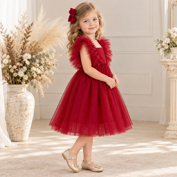A girl in a Callisto red tulle dress with a red bow headband and light shoes stands indoors beside flower arrangements.