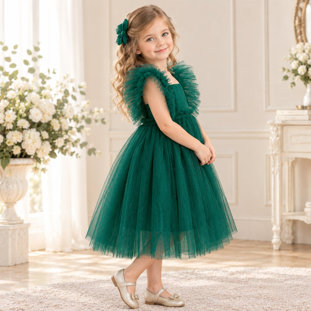 A girl in a Callisto green tulle dress with a red bow headband and light shoes stands indoors beside flower arrangements.