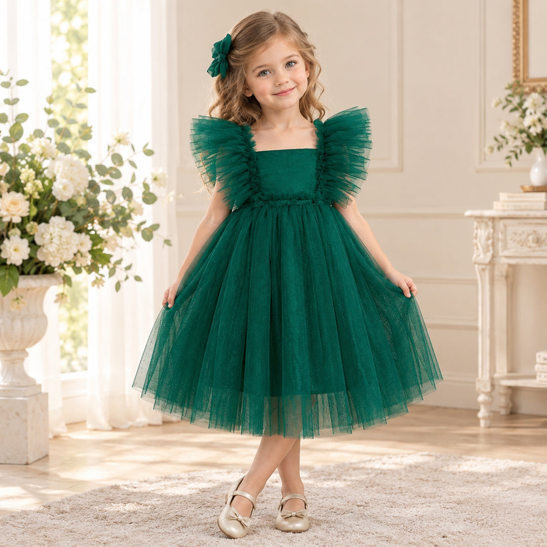 A girl in a Callisto green tulle dress with a red bow headband and light shoes stands indoors beside flower arrangements.