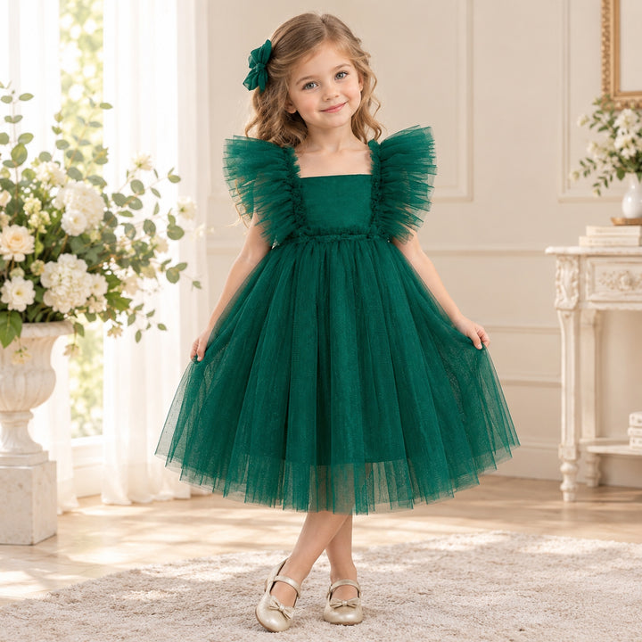 A girl in a Callisto green tulle dress with a red bow headband and light shoes stands indoors beside flower arrangements.