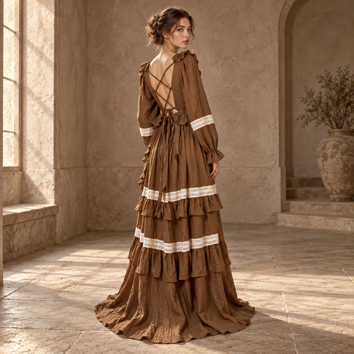 A woman in a Cassandra coffee long-sleeve tiered dress stands with her back turned and head turned, crisscross straps at the back, in a stone interior with a vase on the right.