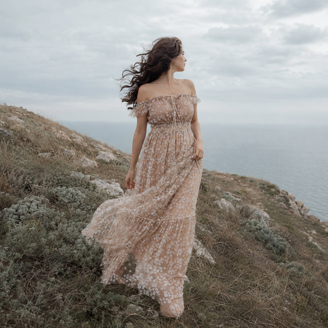A woman stands on a grassy coastal cliff wearing a khaki off-shoulder tulle maxi dress with white floral embroidery, ruffled neckline, and flowing skirt.