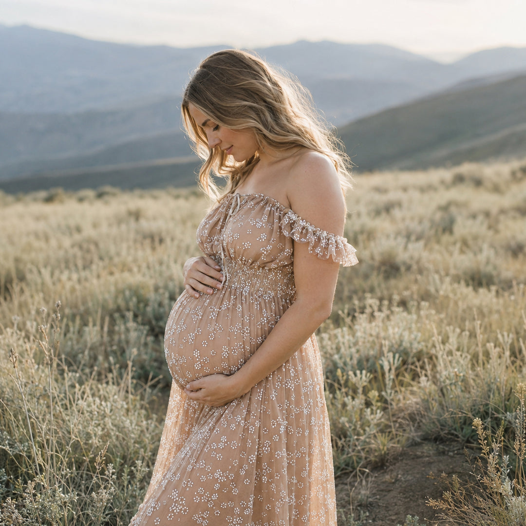 A pregnant woman stands in a grassy mountain landscape wearing a khaki floral embroidered tulle maternity gown with ruffled cap sleeves and a flowing skirt.