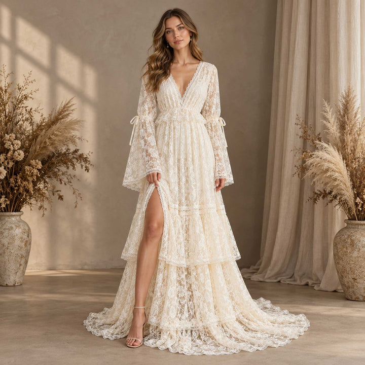 A woman in a Clito beige lace dress with heeled sandals, one hand lifting the slit, indoors with curtains and two dried-flower vases.