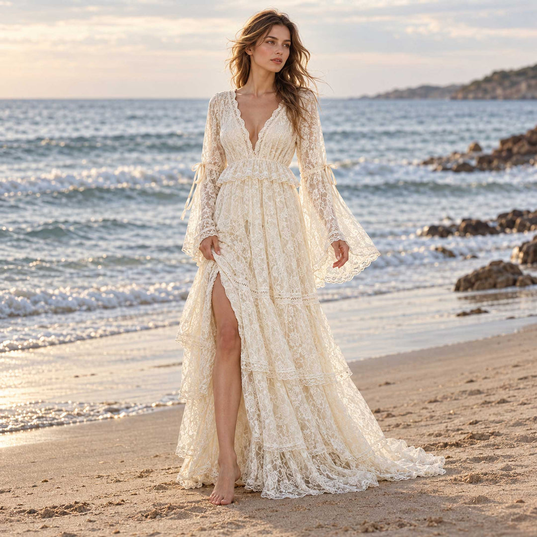 A woman in a Clito beige lace long-sleeve dress with a V neckline, side slit and tiered skirt stands barefoot on a beach, sea and rocks behind.