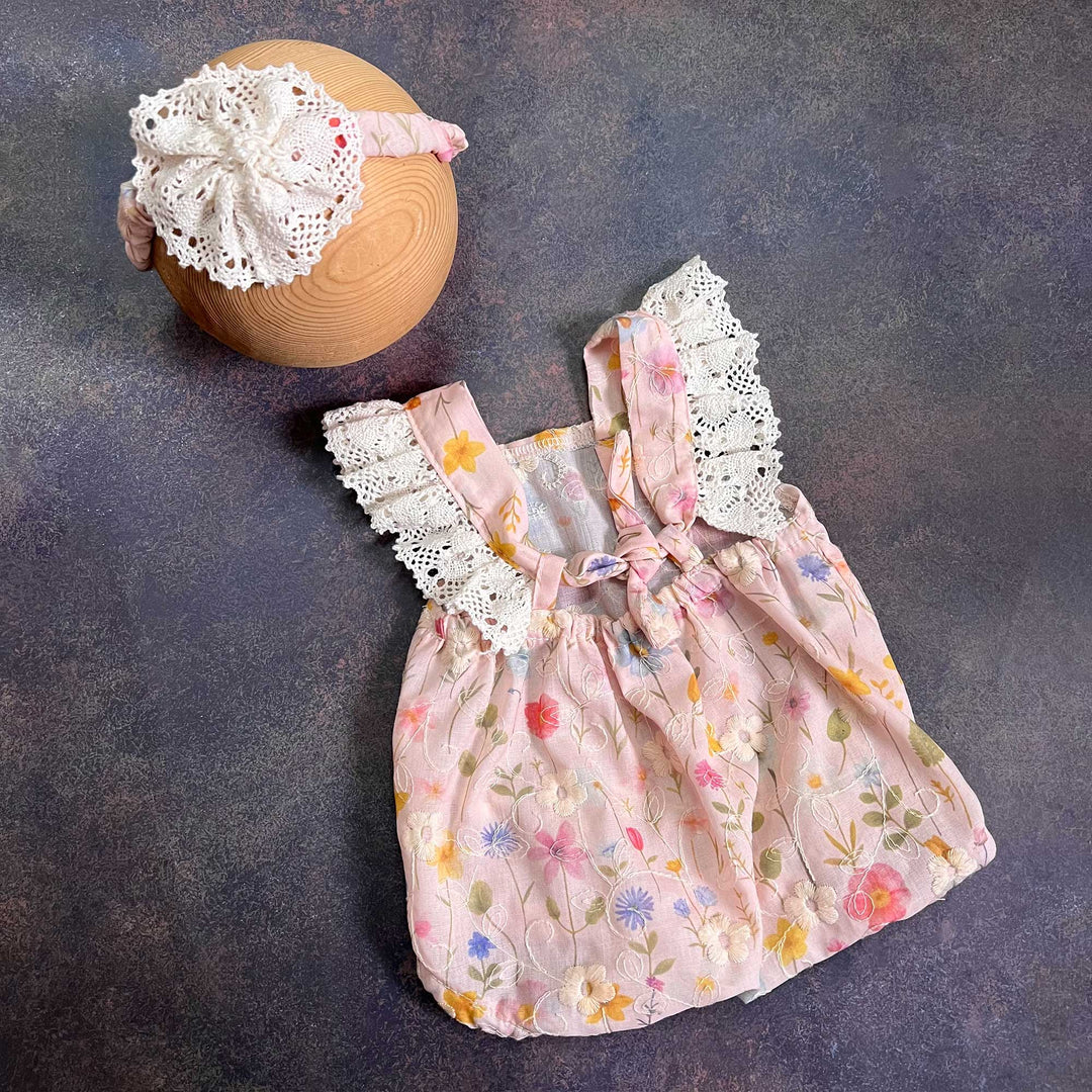 A romper laid flat at an angle, showing the tied back straps, crochet lace and floral fabric, with the same headband at the upper left.