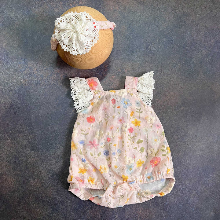A romper laid flat from the back on a dark surface, crochet lace on both straps, the same lace-flower headband placed above.