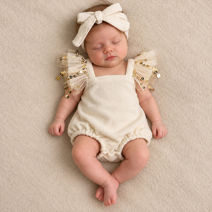 A baby in the same fleece romper and bow headband lies on her back on a beige blanket, eyes closed.
