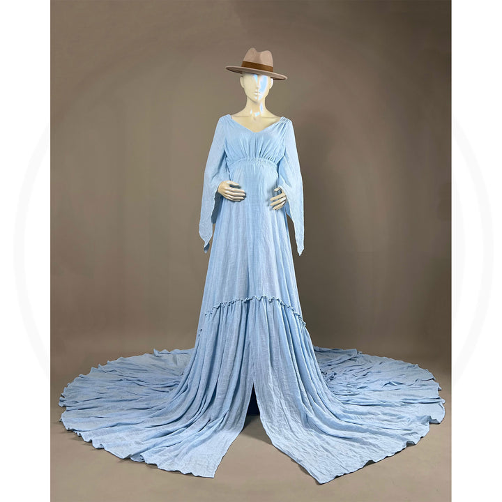 A Daphne light blue long-sleeved V-neck gown is displayed on a mannequin, with hands positioned on the simulated belly. The dress features a high front slit and its hem and train are spread out on the floor.