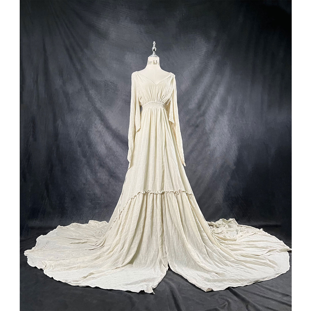A Daphne light grey long-sleeved V-neck gown is displayed on a mannequin, with hands positioned on the simulated belly. The dress features a high front slit and its hem and train are spread out on the wooden floor.

