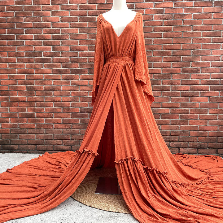 A Daphne rust red long-sleeved V-neck gown is displayed on a mannequin. The dress features a high front slit and its hem and train are spread out on the wooden floor.