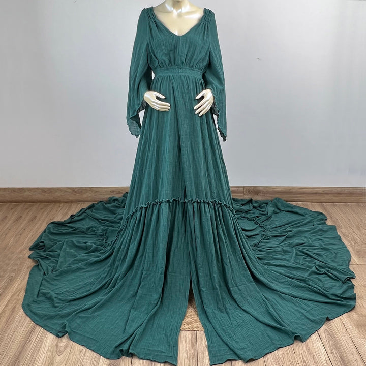 A Daphne emerald long-sleeved V-neck gown is displayed on a mannequin, with hands positioned on the simulated belly. The dress features a high front slit and its hem and train are spread out on the wooden floor.