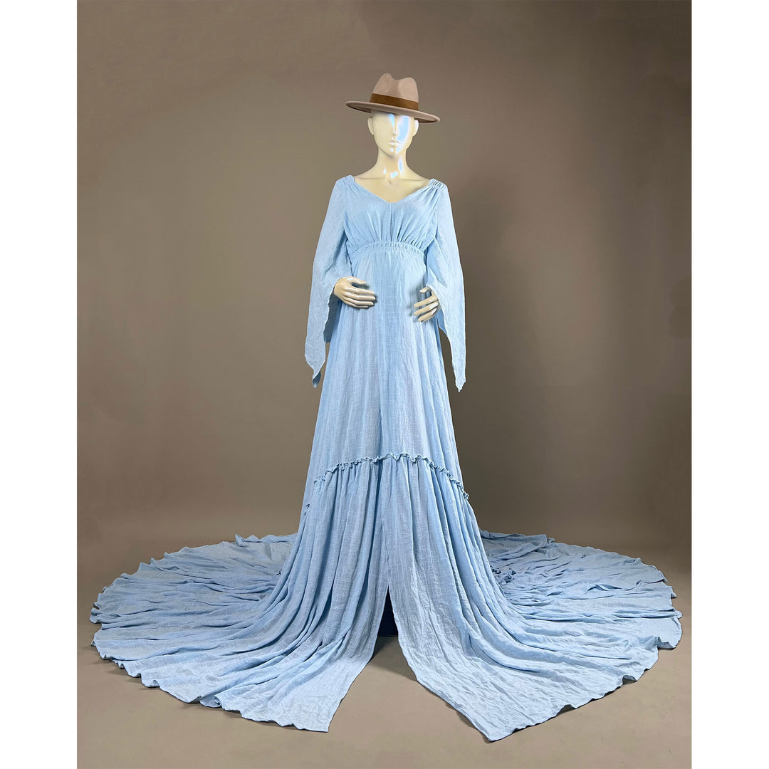 A  Daphne light blue long-sleeved V-neck gown is displayed on a mannequin, with hands positioned on the simulated belly. The dress features a high front slit and its hem and train are spread out on the floor.