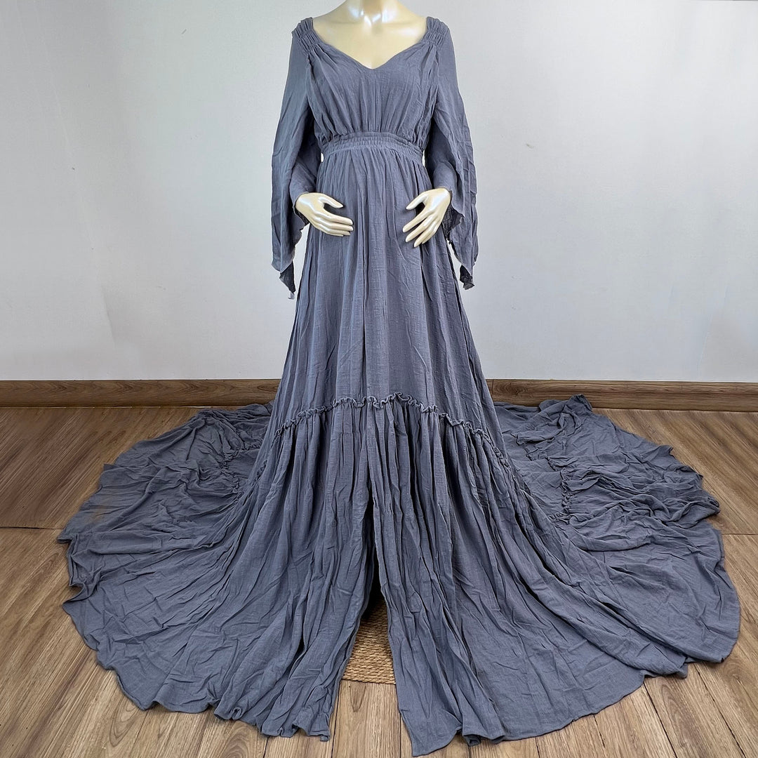 A Daphne blue grey long-sleeved V-neck gown is displayed on a mannequin, with hands positioned on the simulated belly. The dress features a high front slit and its hem and train are spread out on the wooden floor.