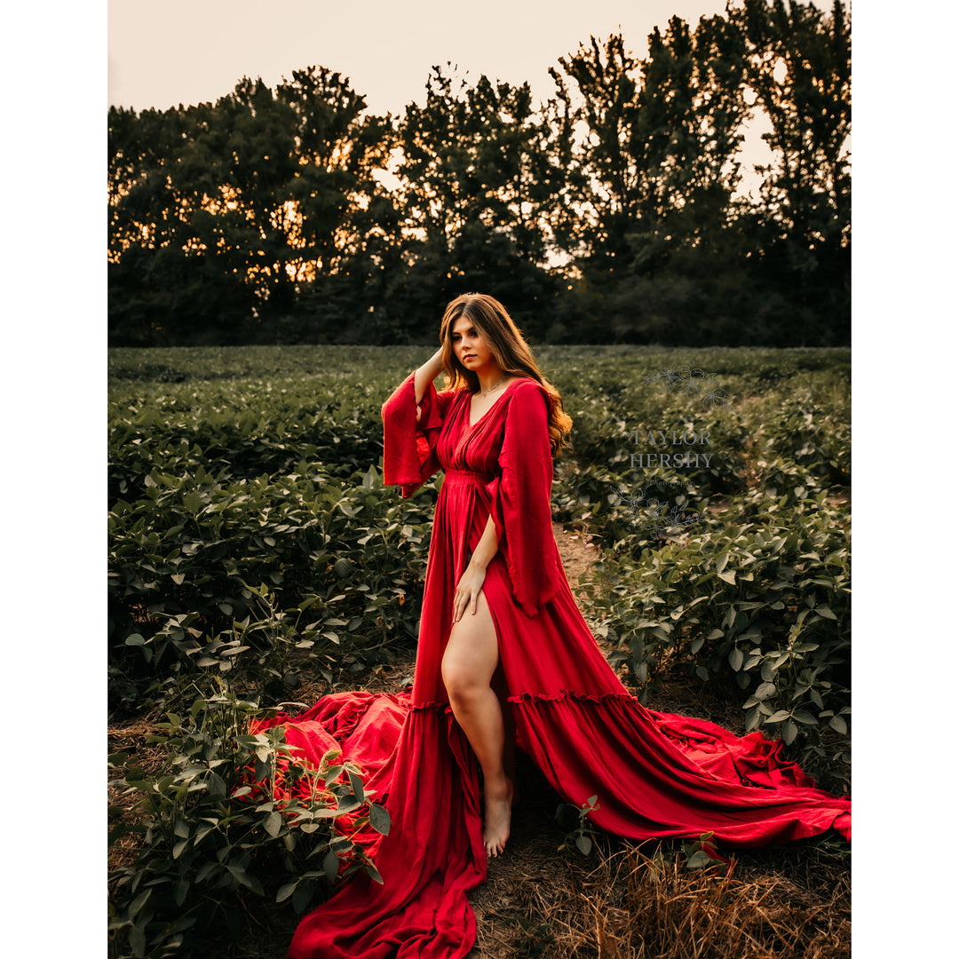 A woman stands in a green crop field wearing a Daphne red gown with sleeves and a side slit. She has one hand on her hair and the other on her thigh.
