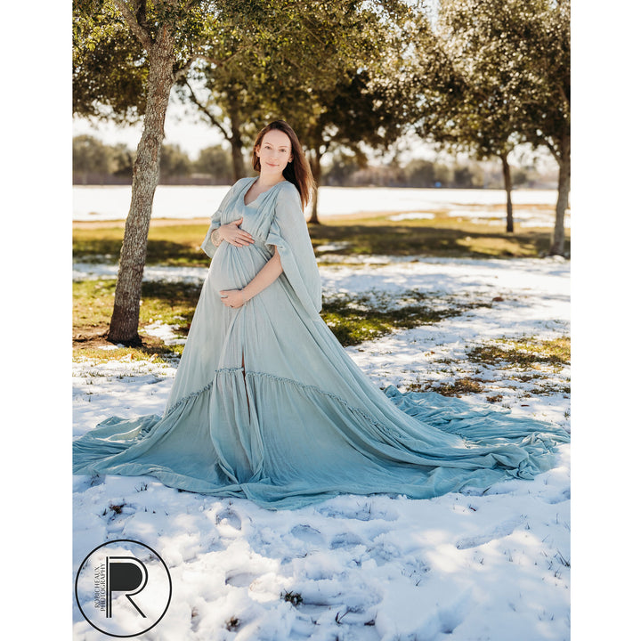 A pregnant woman in a Daphne light blue slit dress with a shawl stands on snow-covered grass, holding her belly.