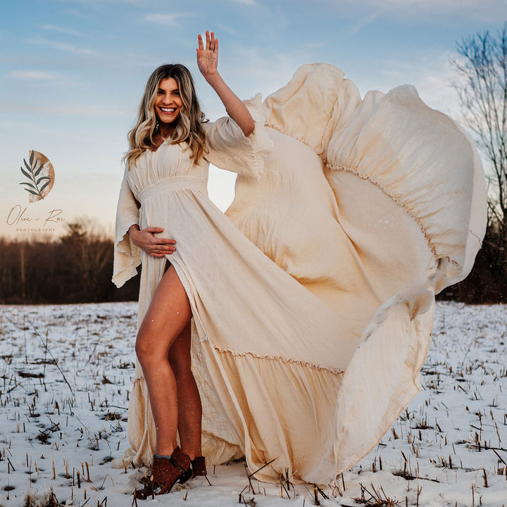 A pregnant woman in a Daphne beige side-slit dress stands in the snow, holding her belly. Her skirt flies up, one hand raised.