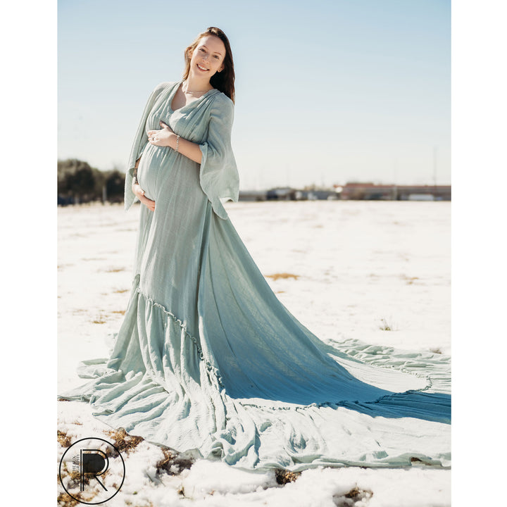 A pregnant woman in a Daphne light blue slit dress with a shawl stands on snow-covered grass, holding her belly.