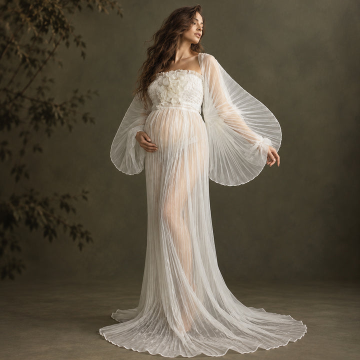 A woman models a floor-length white gown with dramatic sheer billowy sleeves, a textured floral bodice, and a long train against a dark olive-brown studio backdrop.
