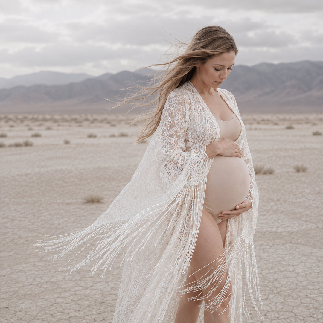 A pregnant woman stands in a dry, cracked desert landscape with mountains in the distance, wearing a beige lace fringe robe over a maternity bodysuit.