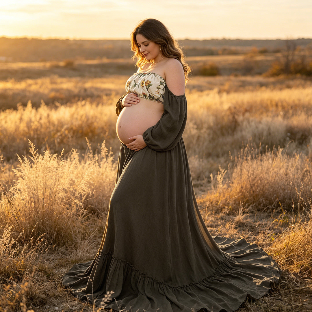 A pregnant woman in a olive gown and floral top stands in tall grass, holding her belly.
