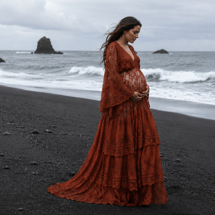 A pregnant woman stands on a black sand beach facing the ocean waves, wearing terracotta lace maternity gown with a deep V-neckline and tiered skirt. 