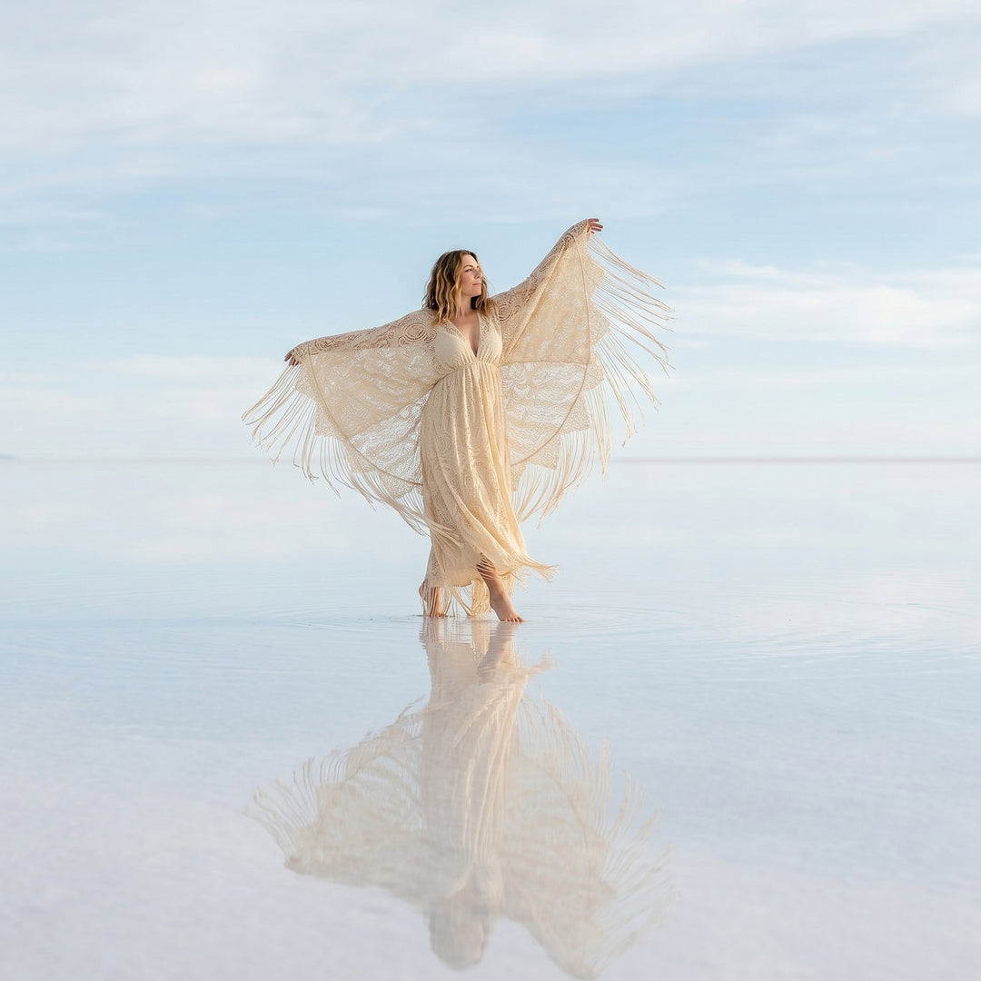 A woman stands in shallow water wearing a beige lace fringe robe with arms spread; the robe features a deep V-neck and wide sleeves.