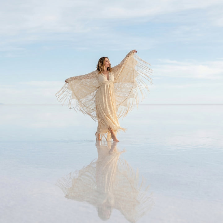 A woman stands in shallow water wearing a beige lace fringe robe with arms spread; the robe features a deep V-neck and wide sleeves.