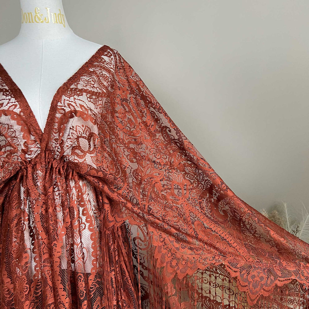 A mannequin displays a close-up of a terracotta lace cape featuring floral openwork texture and a deep V-neck; the fabric is semi-transparent mesh.