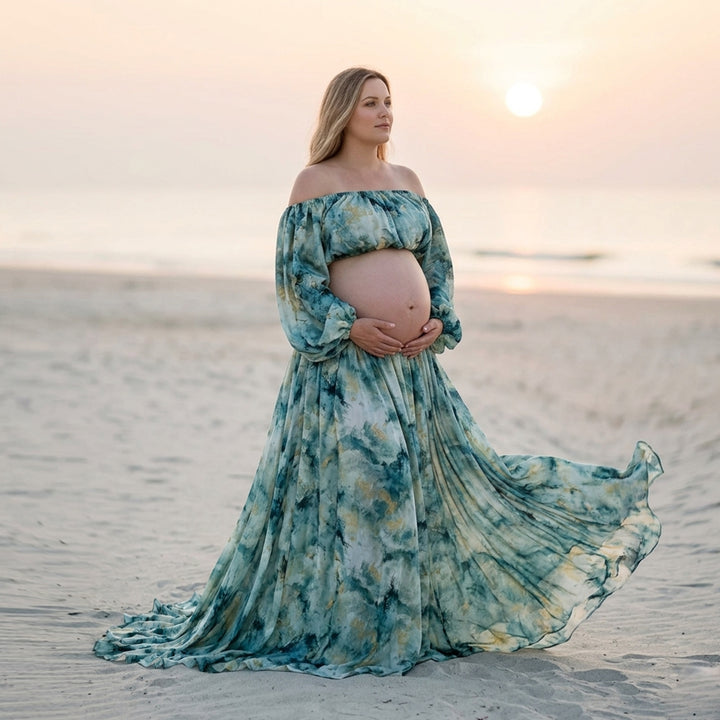 A woman in a Hestia tie dye off shoulder crop top and maxi skirt stands on rocky cliffs by the ocean.holding her belly. 