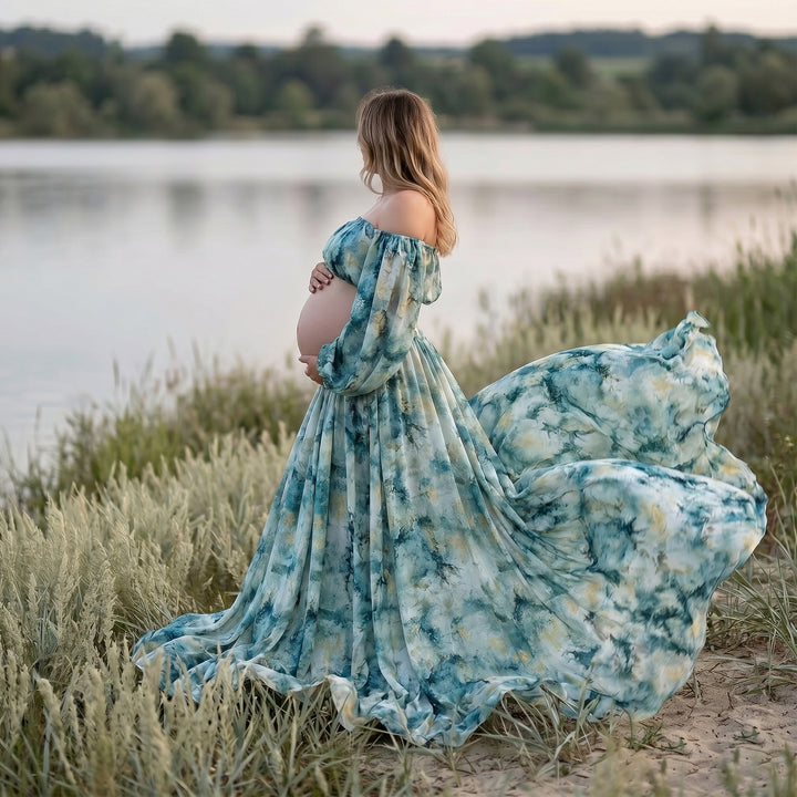 A pregnant woman stands by a lake in a Hestia tie dye print off shoulder dress, holding her belly.