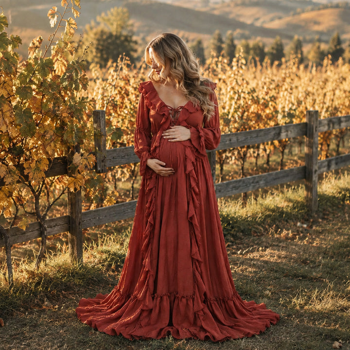 A pregnant woman wearing a long-sleeved terracotta ruffled kallectoria gown stands in a vineyard at sunset, holding her baby bump. 