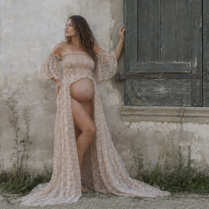 A pregnant woman stands against a rustic textured wall next to an old wooden door, wearing a floral maternity gown with an open front design.