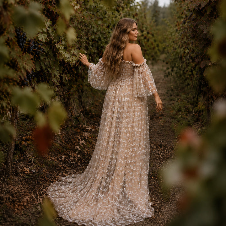 A pregnant woman walks down a path in a vineyard, surrounded by grapevines, wearing a floor-length khaki tulle dress with floral embroidery. 