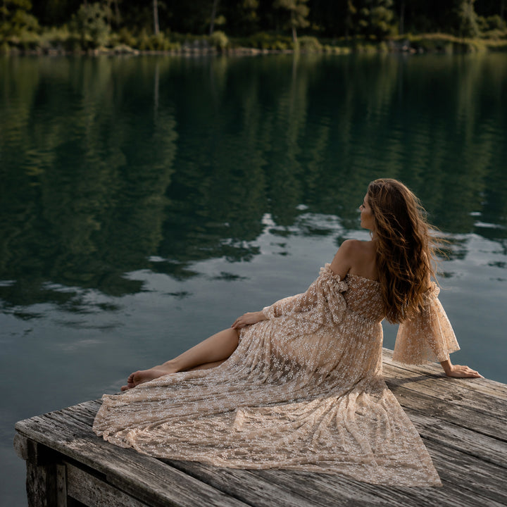 A pregnant woman sits on a weathered wooden dock by a calm lake, wearing a khaki off-shoulder floral tulle gown. 