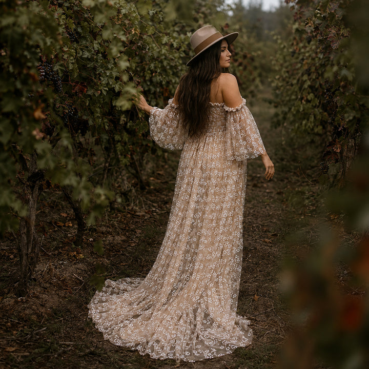 A pregnant woman stands in a vineyard aisle wearing a wide-brimmed hat and a khaki off-shoulder floral gown. 