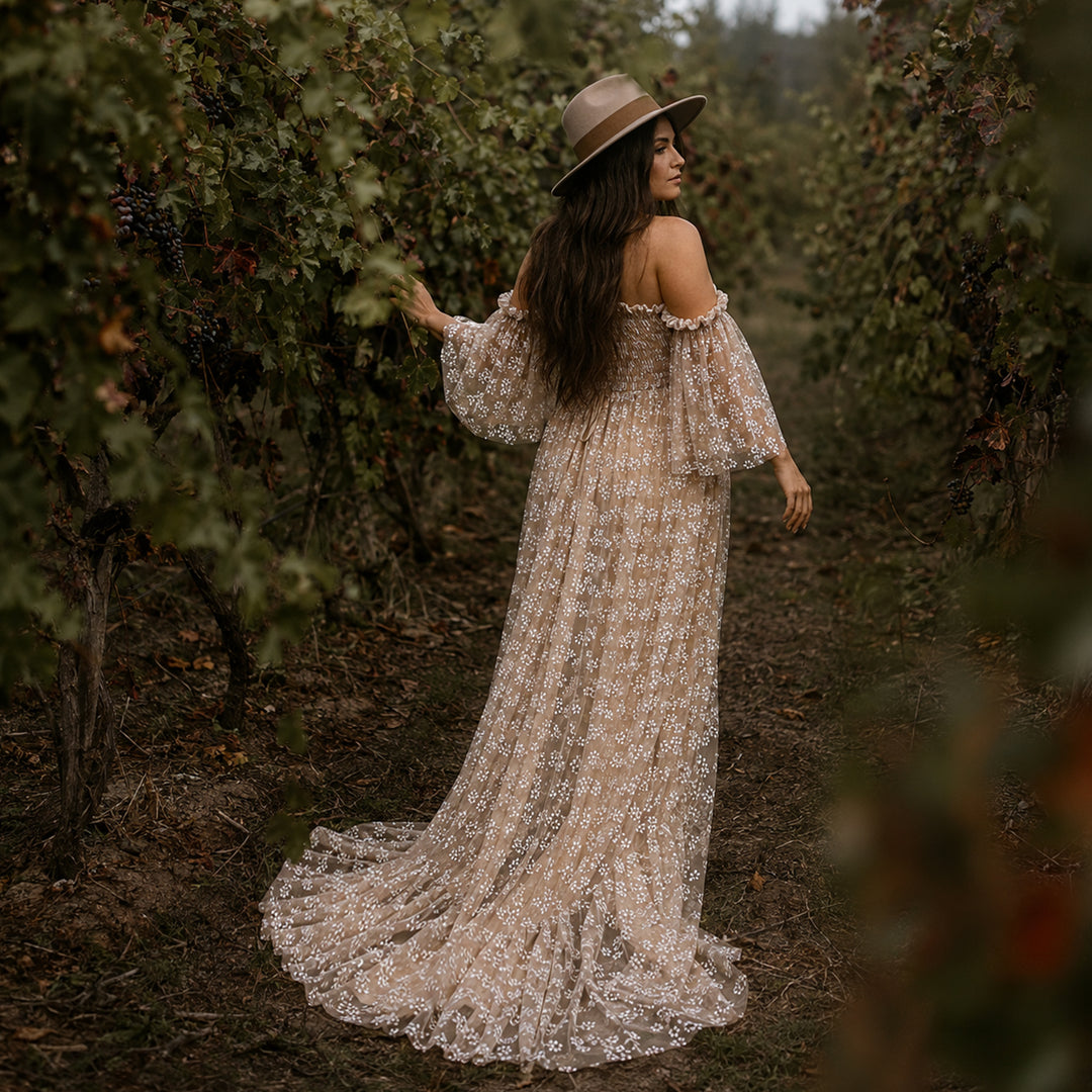 A pregnant woman stands in a vineyard aisle wearing a wide-brimmed hat and a Lysianassa khaiki off-shoulder floral gown. 