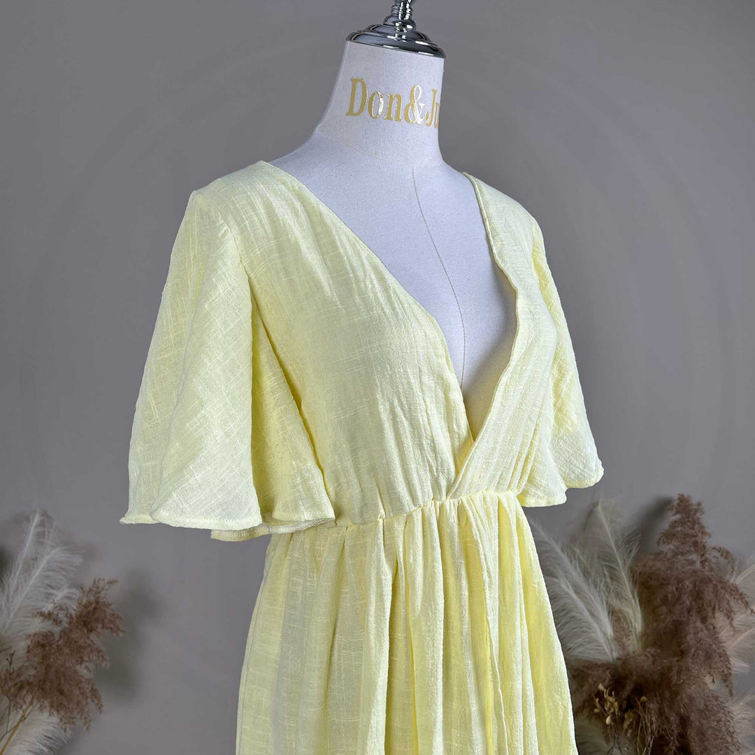 A mannequin displays displays the upper body of yellow V-neck short-sleeve dress, showing a deep-V cut and waist pleats; the fabric has a textured finish, set against a solid wall with dried flowers on both sides.